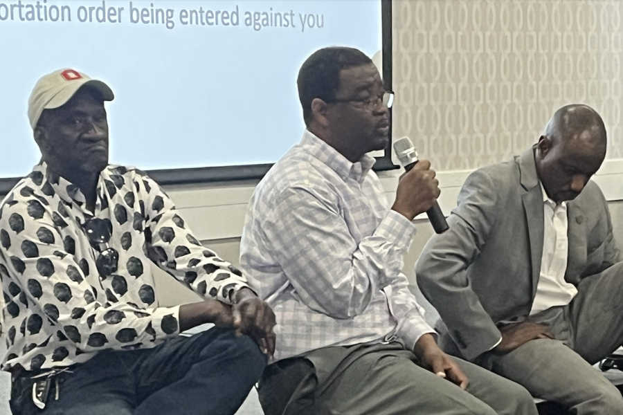 Three Black men sit on a stage and address a community meeting, one holds a mic